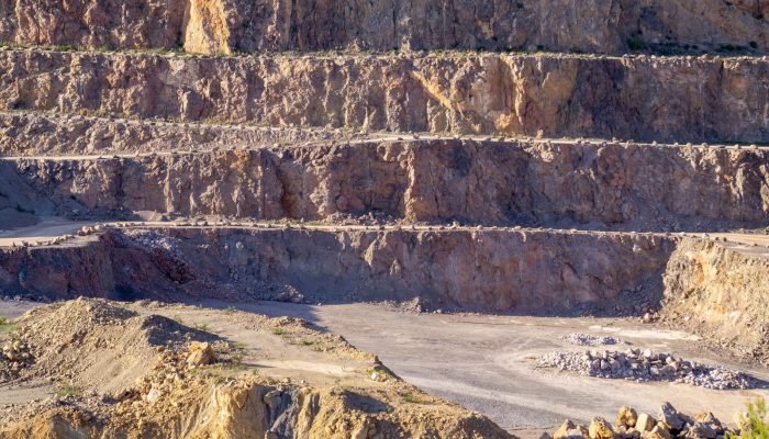 Sunlit view of a rugged, multi-layered rock quarry with exposed strata and rocky terrain.